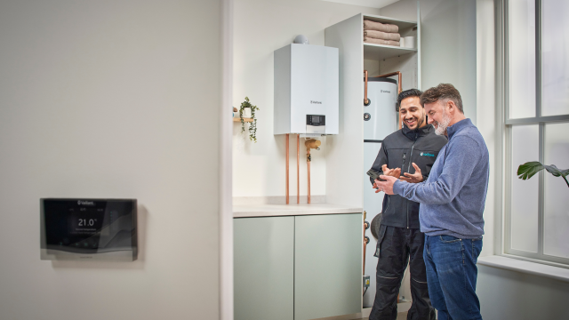 A man and woman stand in a kitchen talking to an installer beside a cylindrical washing machine, engaged in conversation.