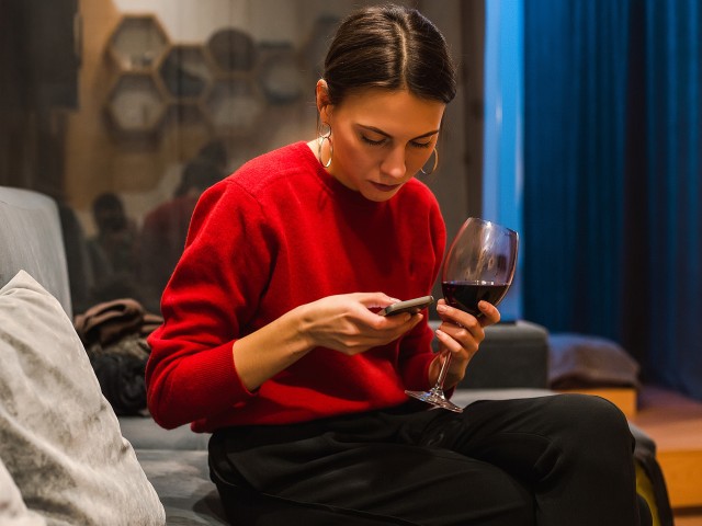 woman in red jumper sat on her phone holding a glass of red wine