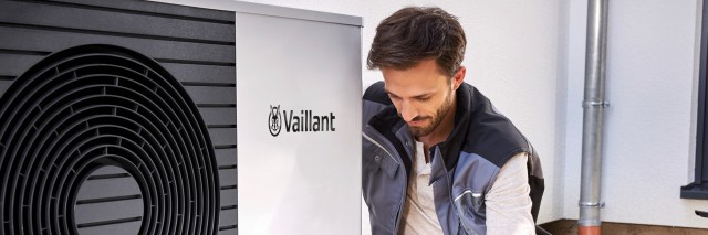 Open kicthen and dining area with a man sat on a sofa watering his plants using a gold watering can. A woman is behind him watching him whilst leaning on the counter. Through the window, you can see a Vaillant heat pump
