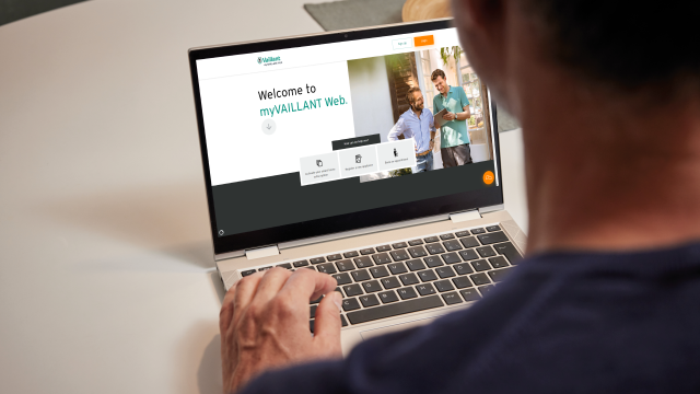 Man sat at desk browsing on laptop. The myVAILLANT Web welcome screen is visible