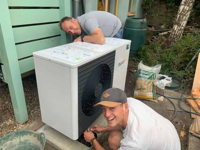 a heat pump in the middle with one installer bending down wearing a hat and the other installer leaning over to the top of the heat pump