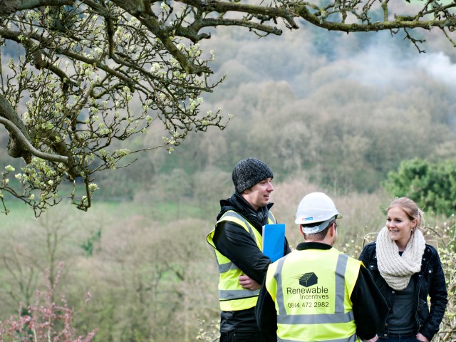 three people standing in a field with trees surrounding them. Two men are wearing a high vis jacket