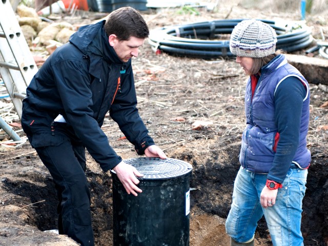 an installer holding a black cylinder into the ground with a woman looking at where the cylinder is being placed. There are two black pipes coming out at the bottom of the cylinder