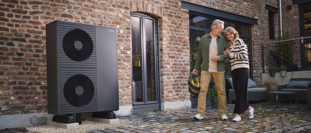 2 air-source heat pumps outside a big red brick home and a couple walking by