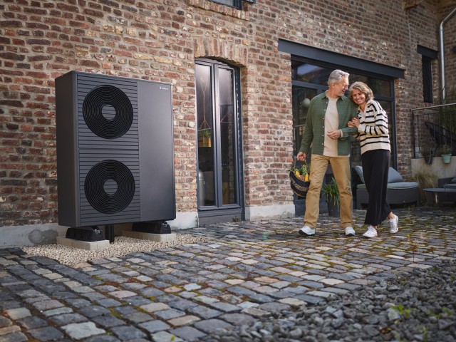 2 air-source heat pumps outside a big red brick home and a couple walking by