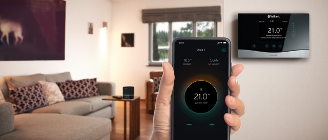 hand holding an iPHONE with a thermostat on the wall. The background is blurred and you can see a sofa, Amazon ALEXa 