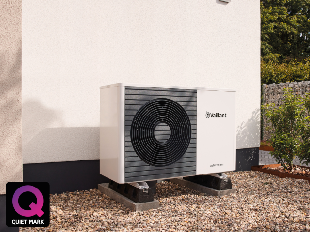 aroTHERM plus heat pump outside a wall on a white background and bushes. The quiet mark logo sits on the bottom left corner
