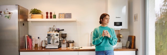 woman holding a mug looking outside a window with a boiler to the right and other kitchen items in the background