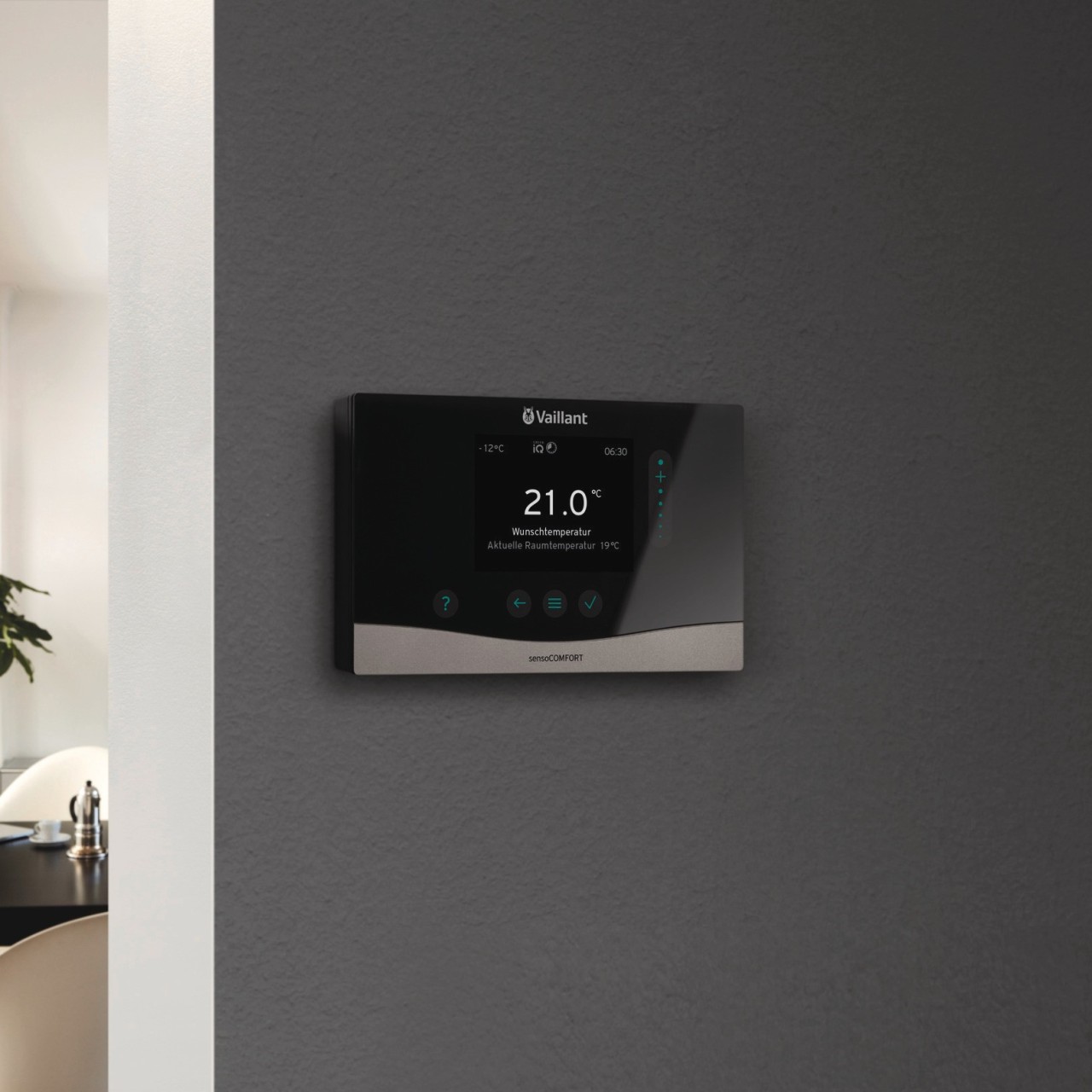 sensoCOMFORT thermostat on a dark grey wall with soft furnishing to the left with a table in the background.