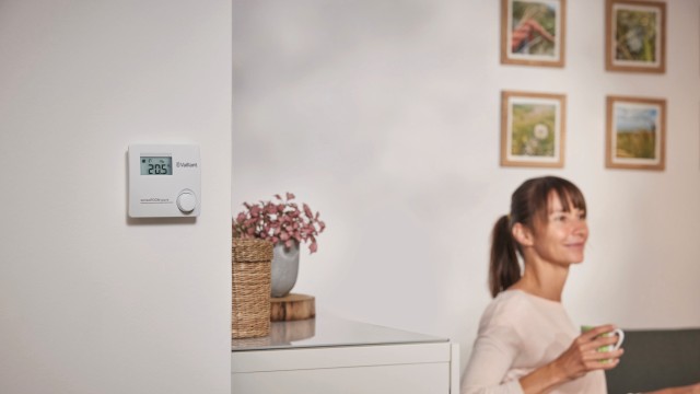 a sensoROOM pure heating control on a wall, a woman is sat on the sofa in the background with a mug in her hand.