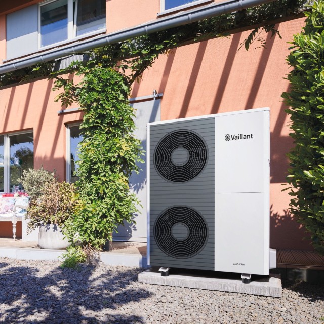 double fan aroTHERM plus heat pump outside a red wall with leaves and bushes