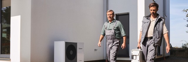 two men installers walking outside, one is holding a tool box. There is a Vaillant air source heat pump in the background.