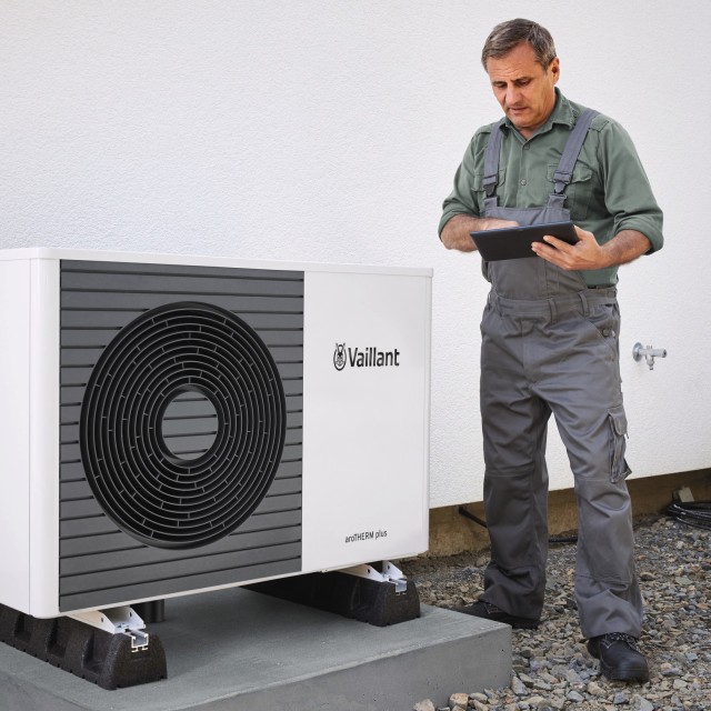 a male installer holding a clipboard with a Vaillant arotherm plus heat pump to the left of him. The heat pump is outside in front a white wall