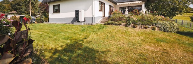 a white bungalow with a grassy garden and an aroTHERM plus heat pump in front of it. There is blue skies and clouds