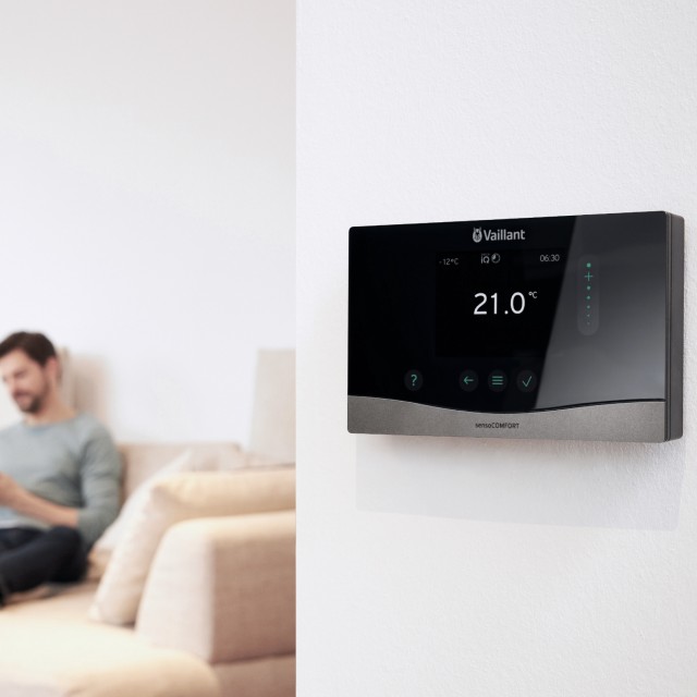 a man sat on his sofa looking at his phone with a Vaillant sensocomfort heating control on a wall