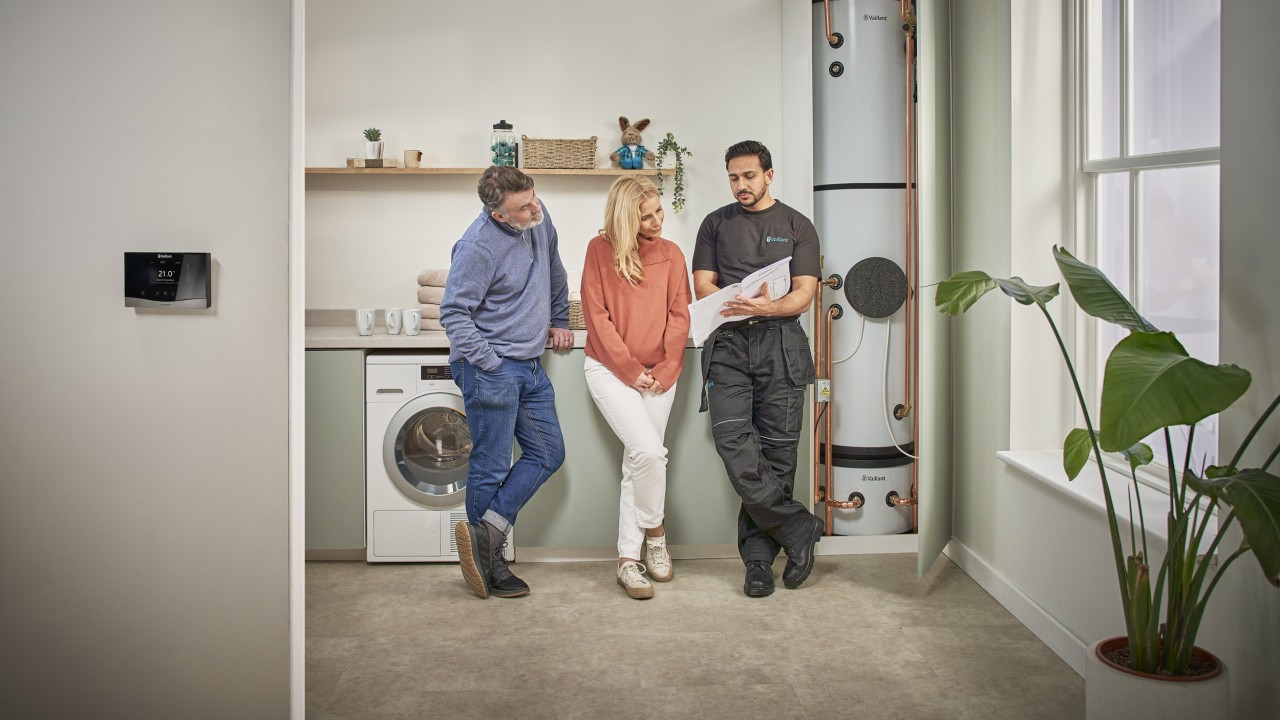 A man and woman stand in a kitchen talking to an installer beside a cylindrical washing machine, engaged in conversation.