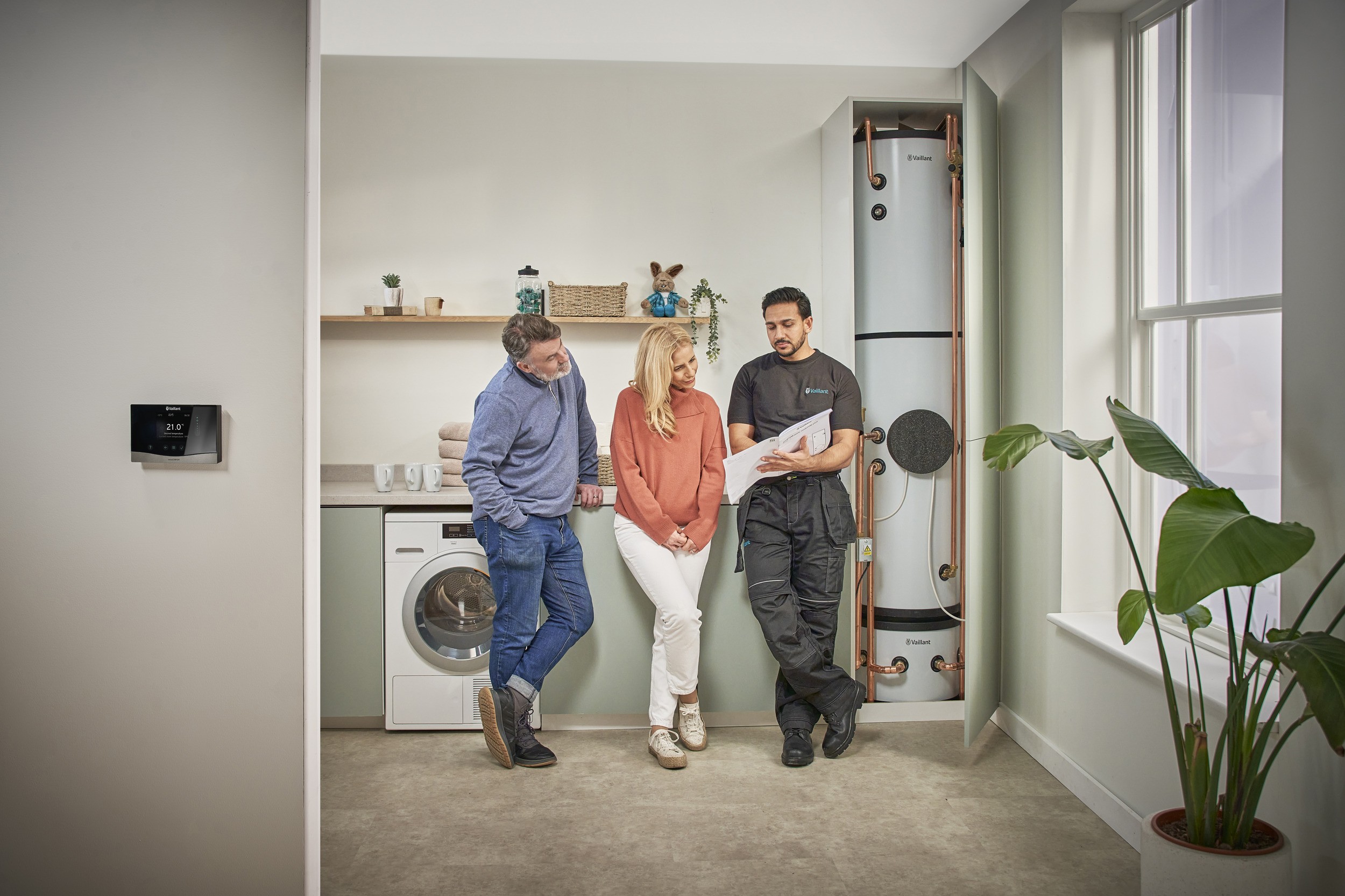 A man and woman stand in a kitchen talking to an installer beside a cylindrical washing machine, engaged in conversation.